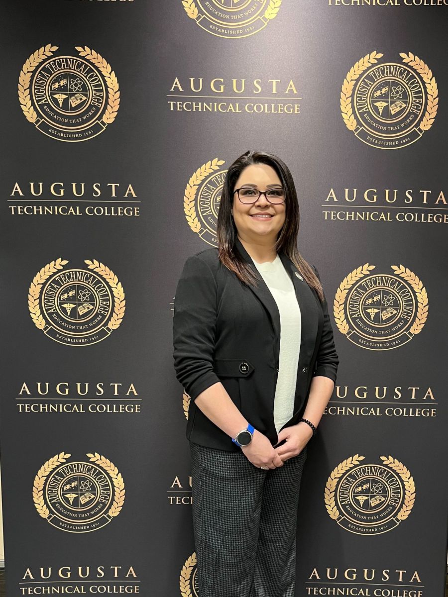 Devlynn Taylor, a caucasian female, stands in front of a backdrop with the Augusta Technical College seal and name in black and gold. She is wearing a black blazer with a white crew neck dress shirt and dark grey dress pants.