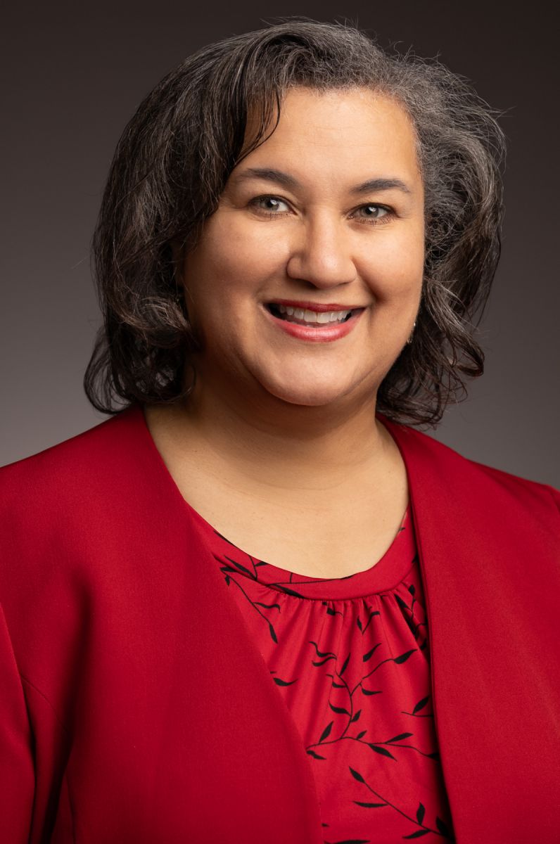 Dr. Melissa Frank-Alston, a Caucasian female with short brown hair streaked grey, smiles wearing a red dress shirt with a black vine print and a red dress jacket pictured against a grey backdrop.