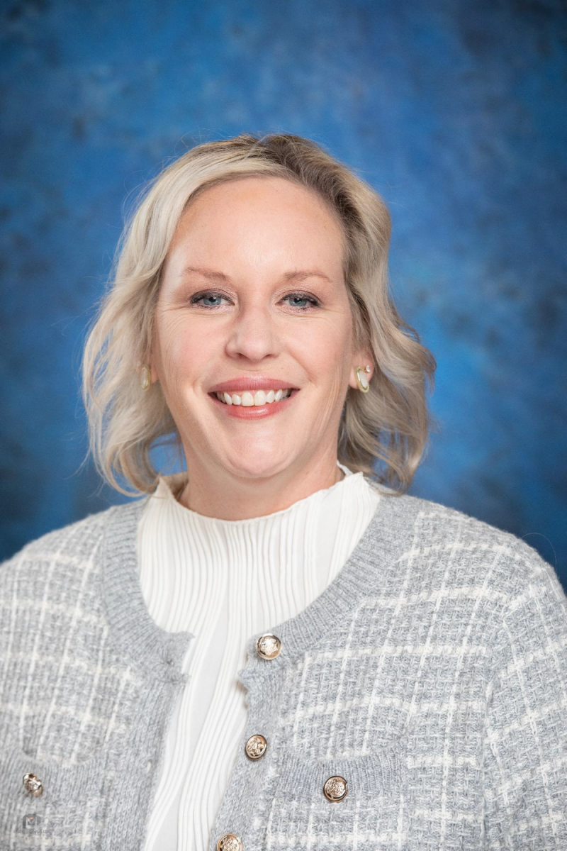 Dr. Beth Sacksteder, a Caucasian female with shoulder length blonde hair smiles at the camera wearing a white sweater with a white plaid cardigan pictured against a blue backdrop.