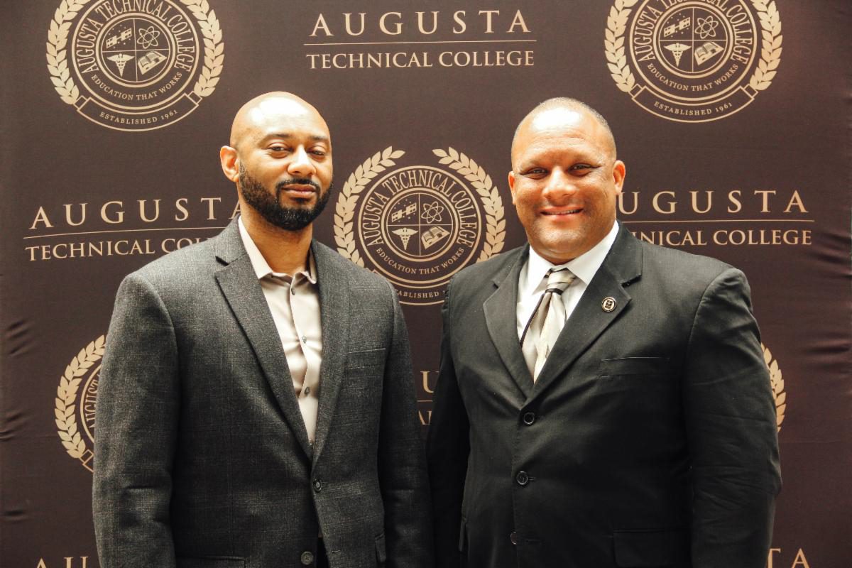 An African American male wearing a grey suit with a white shirt and a Caucasian male also wearing a grey suit with a white shirt and tie stand in front of a background with the words Augusta Technical College and the Augusta Technical College seal in gold on a black background.