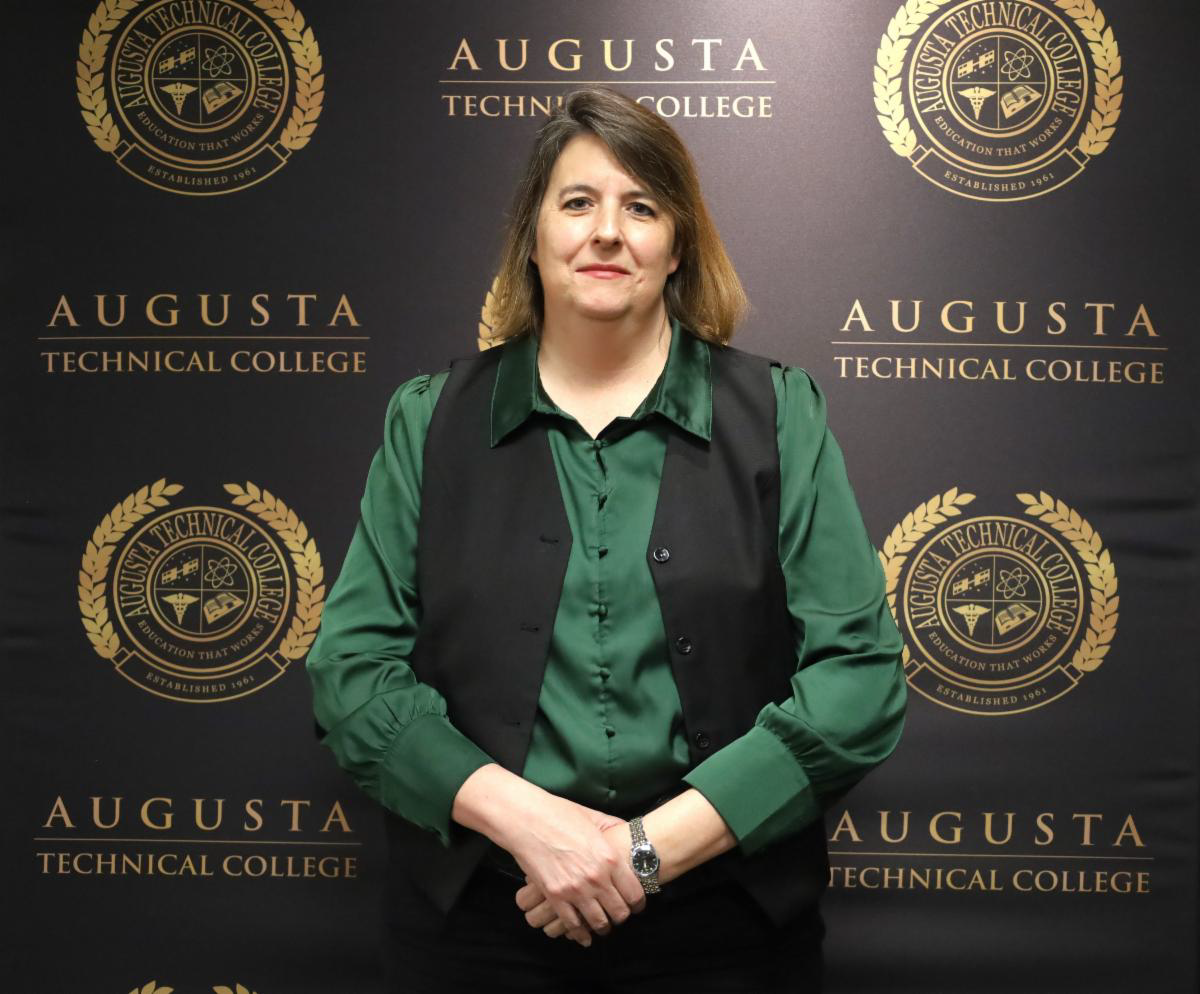 Chief Jerri Jennings-Joly, a Caucasian female with shoulder length dark blonde hair, smiles at the camera wearing a green collared dress shirt under an open black vest with black trousers. She stands against a black backdrop alternating the gold Augusta Technical College seal and the words Augusta separated by a horizontal line from the words Technical College also in gold.