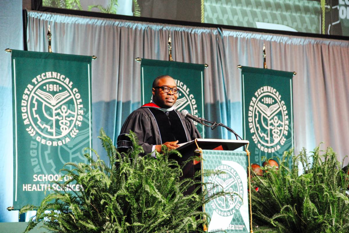 Dr. Whirl wearing commencement regalia stands at podium giving speech with the school banners in the background and ferns on the left and right side of the podium.