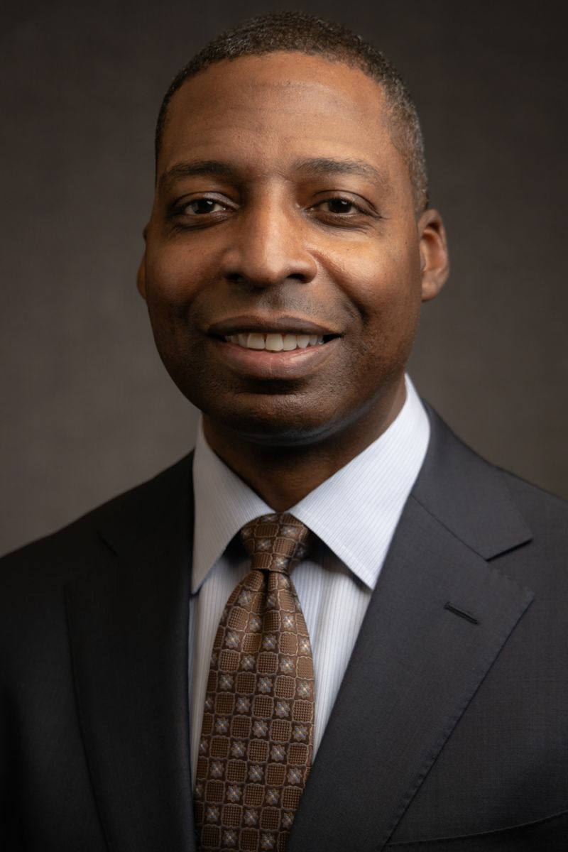 Mr. James Heffner, an African American male, smiles at the camera wearing a charcoal suit jacket with a white collared shirt and brown tie with tan polka dots.