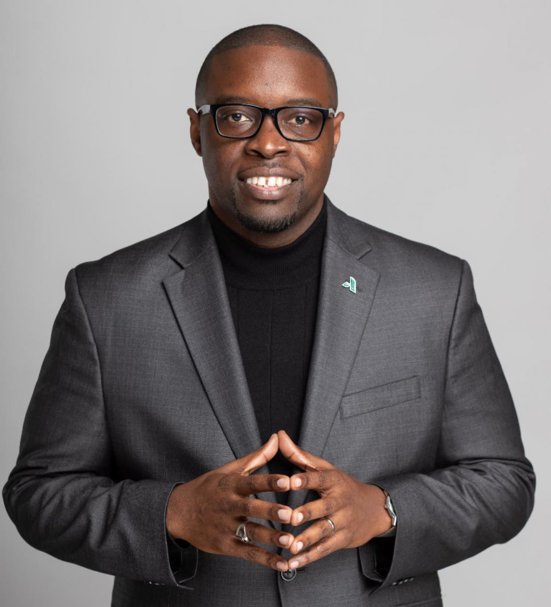 Dr. Jermaine Whirl, an African American male, smiles wearing a grey suit jacket over a black shirt with his hands steepled towards the camera in front of his abdomen.