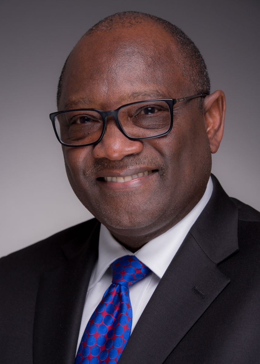 Mr. John Richardson, an African American male, smiling wearing black frame glasses, a black suite with a blue tie with a red pattern.