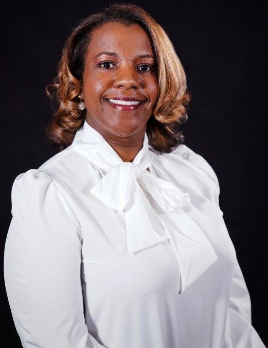 An African American female, Dr. Kristie Searcy, Dean of Health Sciences, has medium length blonde curly hair and smiles wearing a white dress shirt with a bow neck and pearl earrings against a black backdrop.