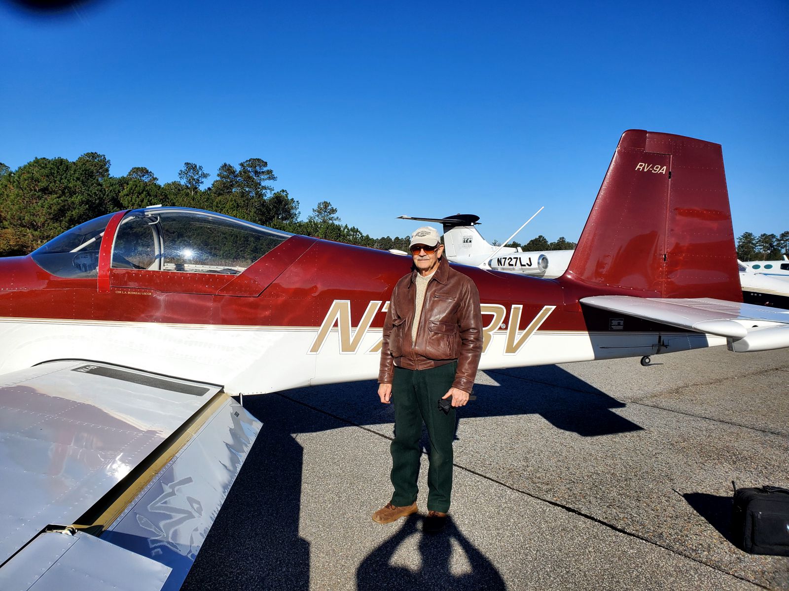 Lawrence Budreau, an older Caucasian male, stands in front of a kit aircraft wearing a brown leather jacket, black pants, brown boots and a tan hat.