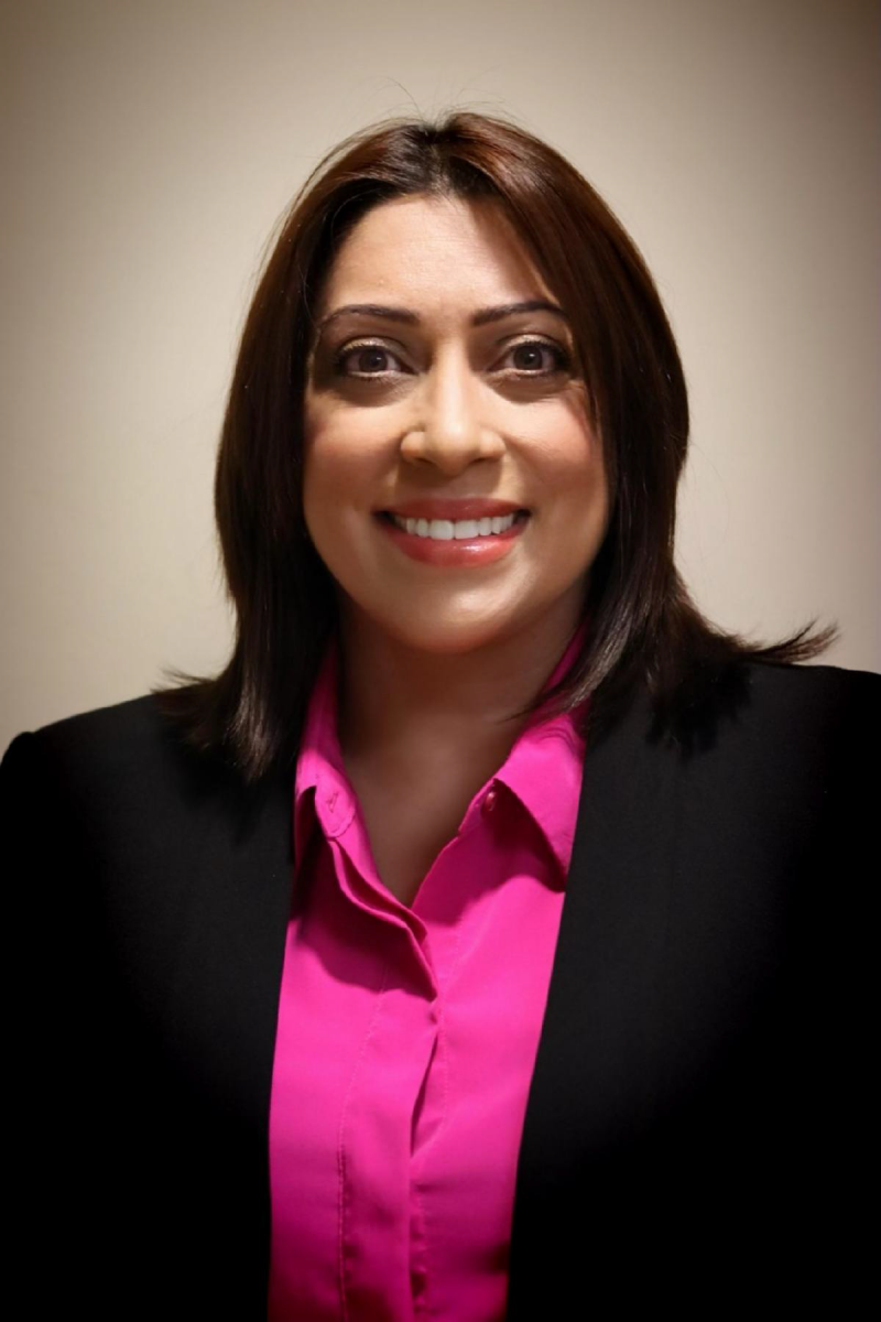 Ms. Migdaliz Berrios, a Latino female, smiles at the camera wearing a black suit jacket with a fuschia collared shirt pictured against a tan background.