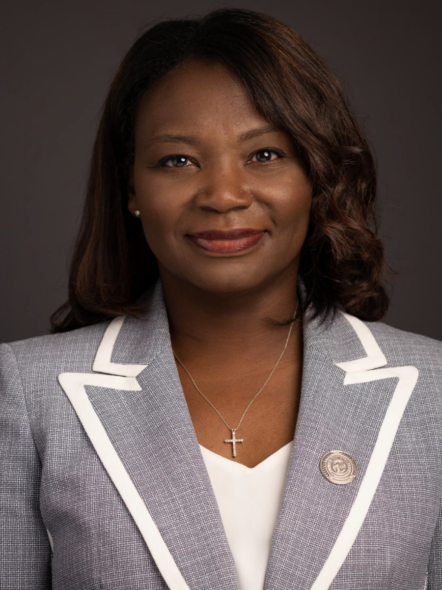 Tara Jenkins, an African American female with shoulder length wavy brown hair, smiles wearing a grey suit jacket with white bordering, a white dress shirt and a gold cross necklace.